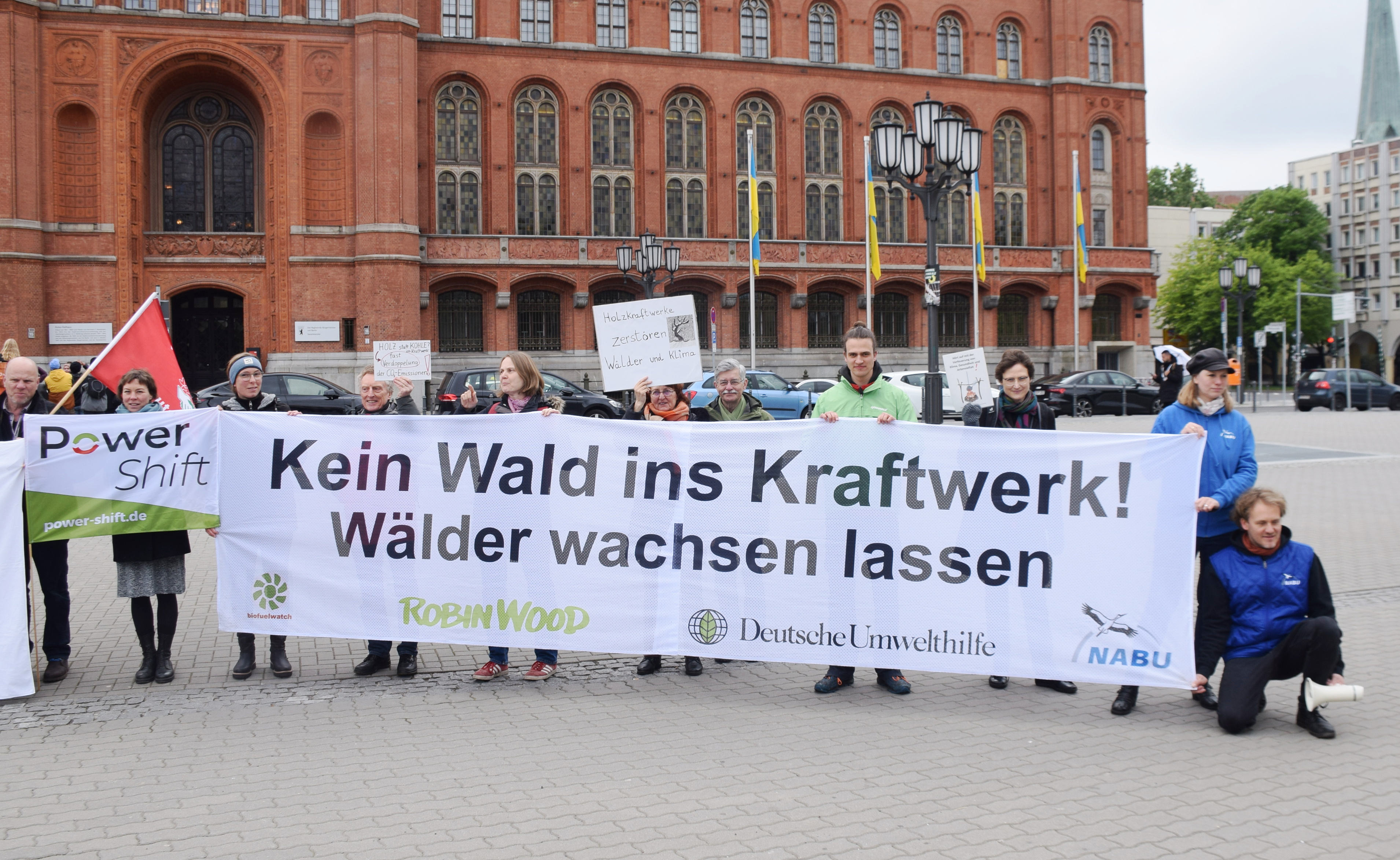 Photo of people outside the red city hall in Berlin, holding placards and a banner with the logos of Biofuelwatch, ROBIN WOOD, Deutsche Umwelthilfe and NABU, saying "Kein Wald ins Kraftwerk! Wälder wachsent lassen" 